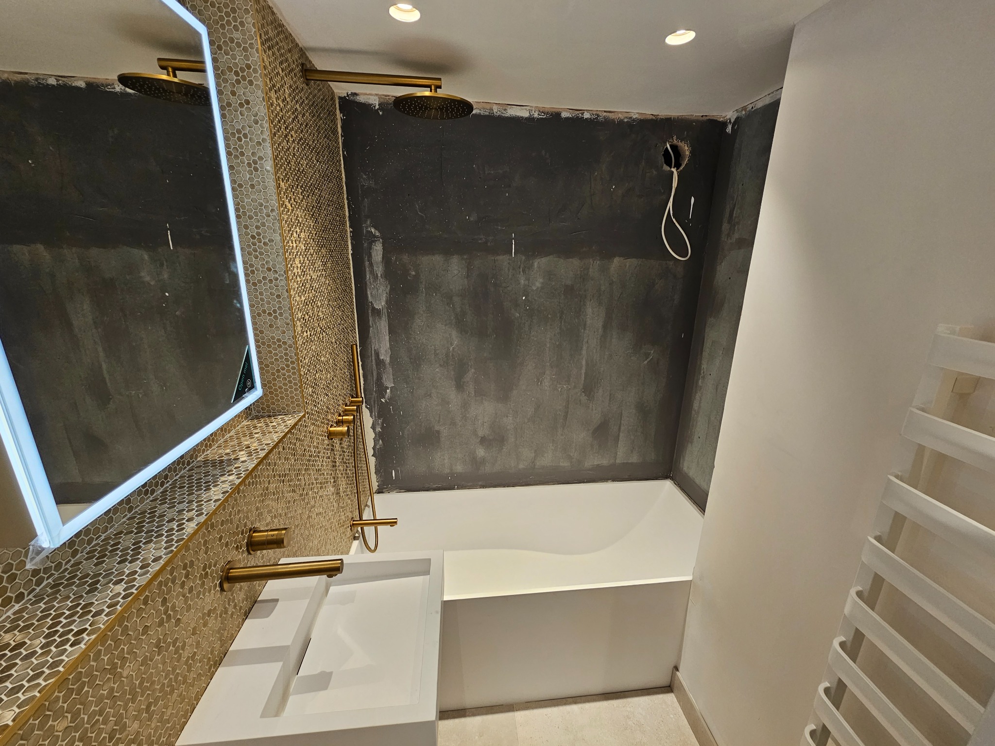 Łazienka w trakcie remontu: biała wanna i umywalka, złota armatura, mozaika w odcieniach złota, ściany częściowo przygotowane pod tynk, widoczny grzejnik drabinkowy.