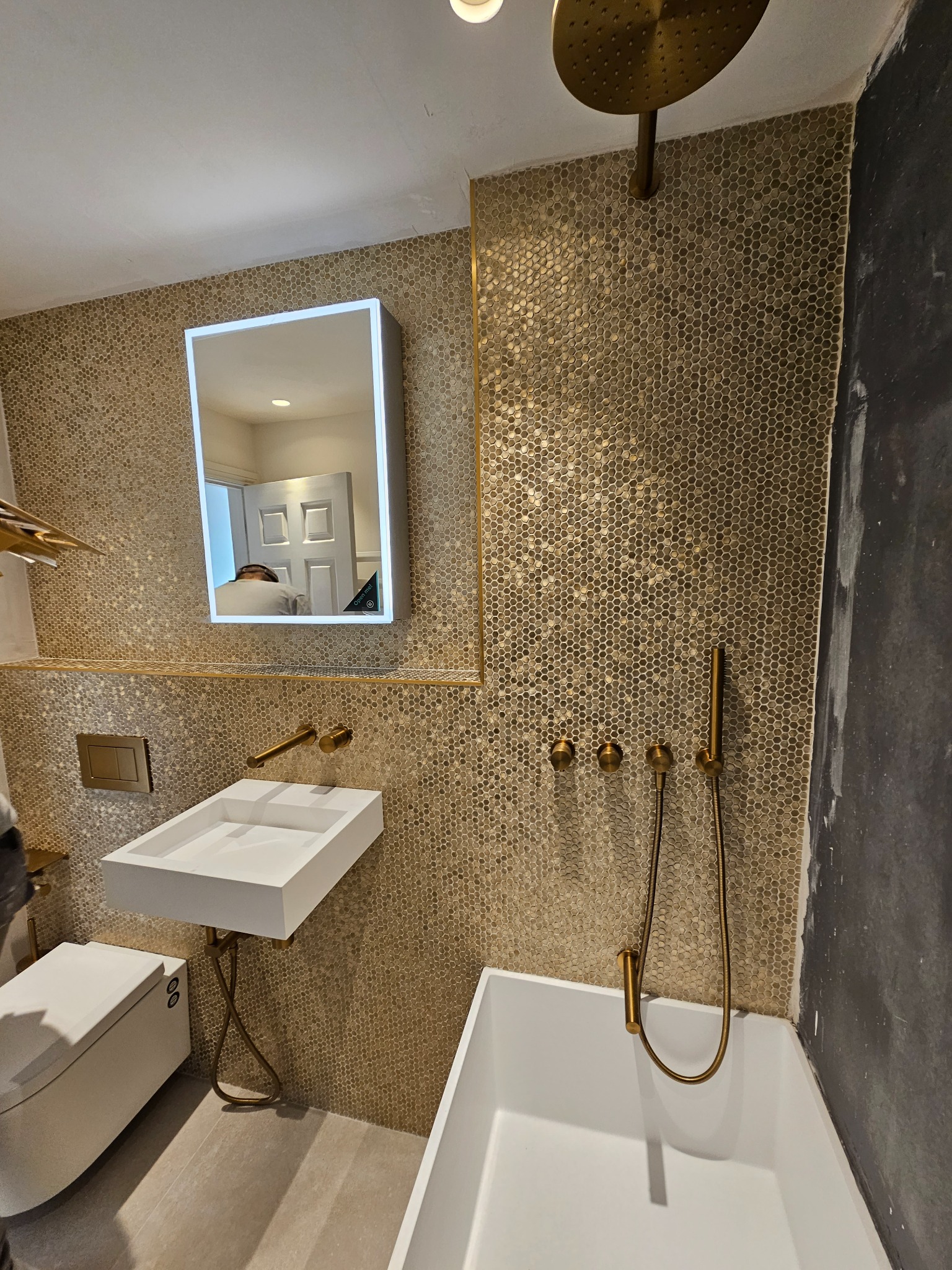 Nowoczesna łazienka z mozaiką w odcieniach złota, białą umywalką i wanną, armaturą w kolorze mosiądzu oraz podświetlanym lustrem, w którego odbiciu widać fragment wnętrza.