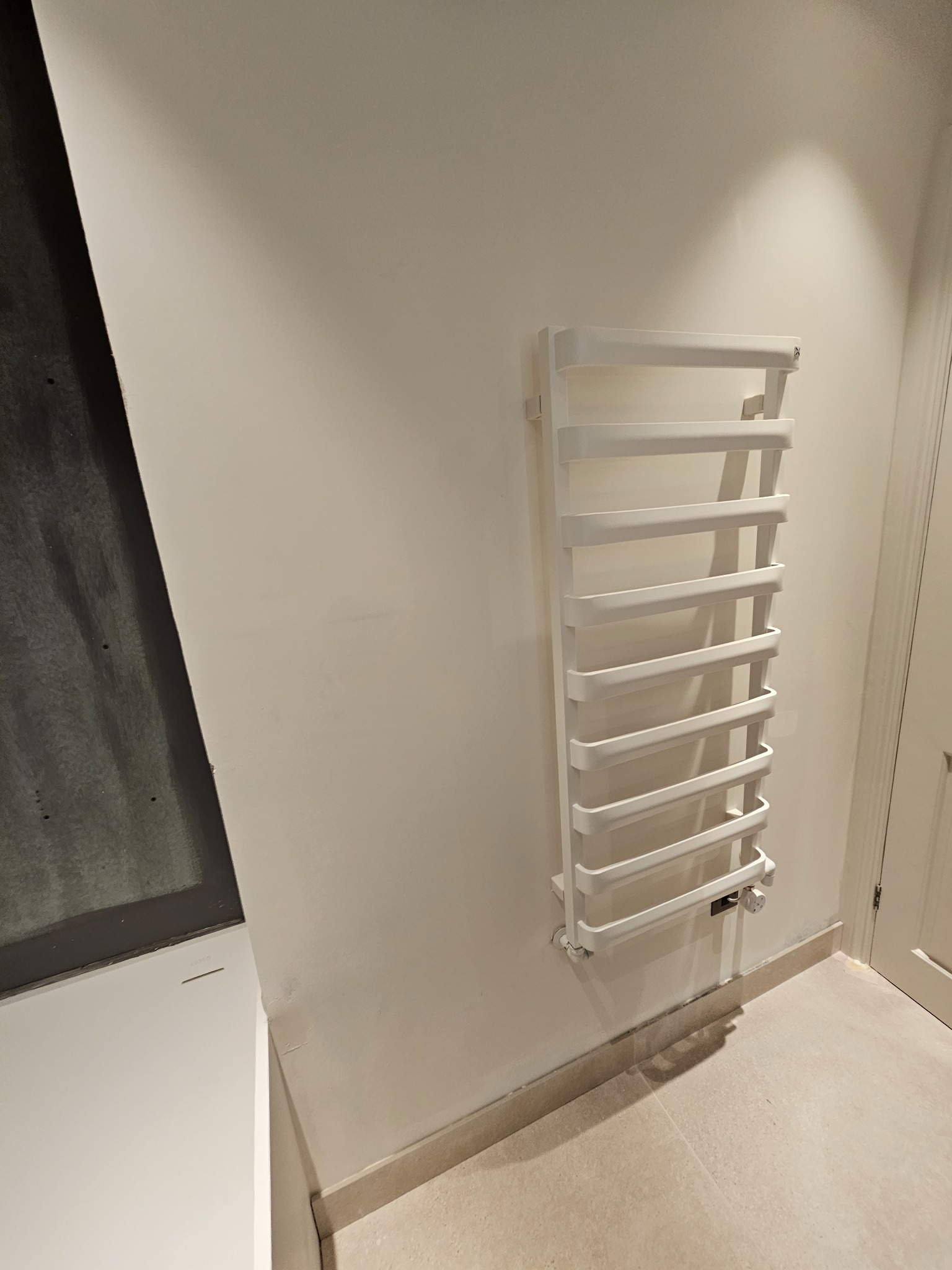 Biały, drabinkowy grzejnik łazienkowy zamontowany na jasnej ścianie, z widocznym termostatem, na tle beżowej podłogi i fragmentu drzwi.