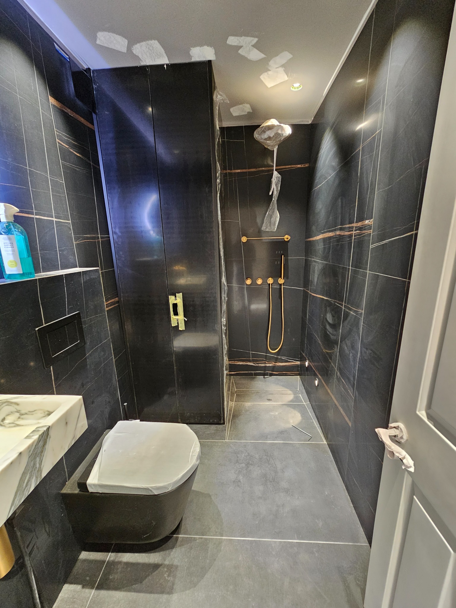 Wykończona łazienka z czarnymi płytkami z wyraźnym złotym żyłowaniem, wisząca muszla klozetowa i armatura prysznicowa w kolorze złotym, zabezpieczone folią ochronną, widoczne ślady po gipsowaniu...