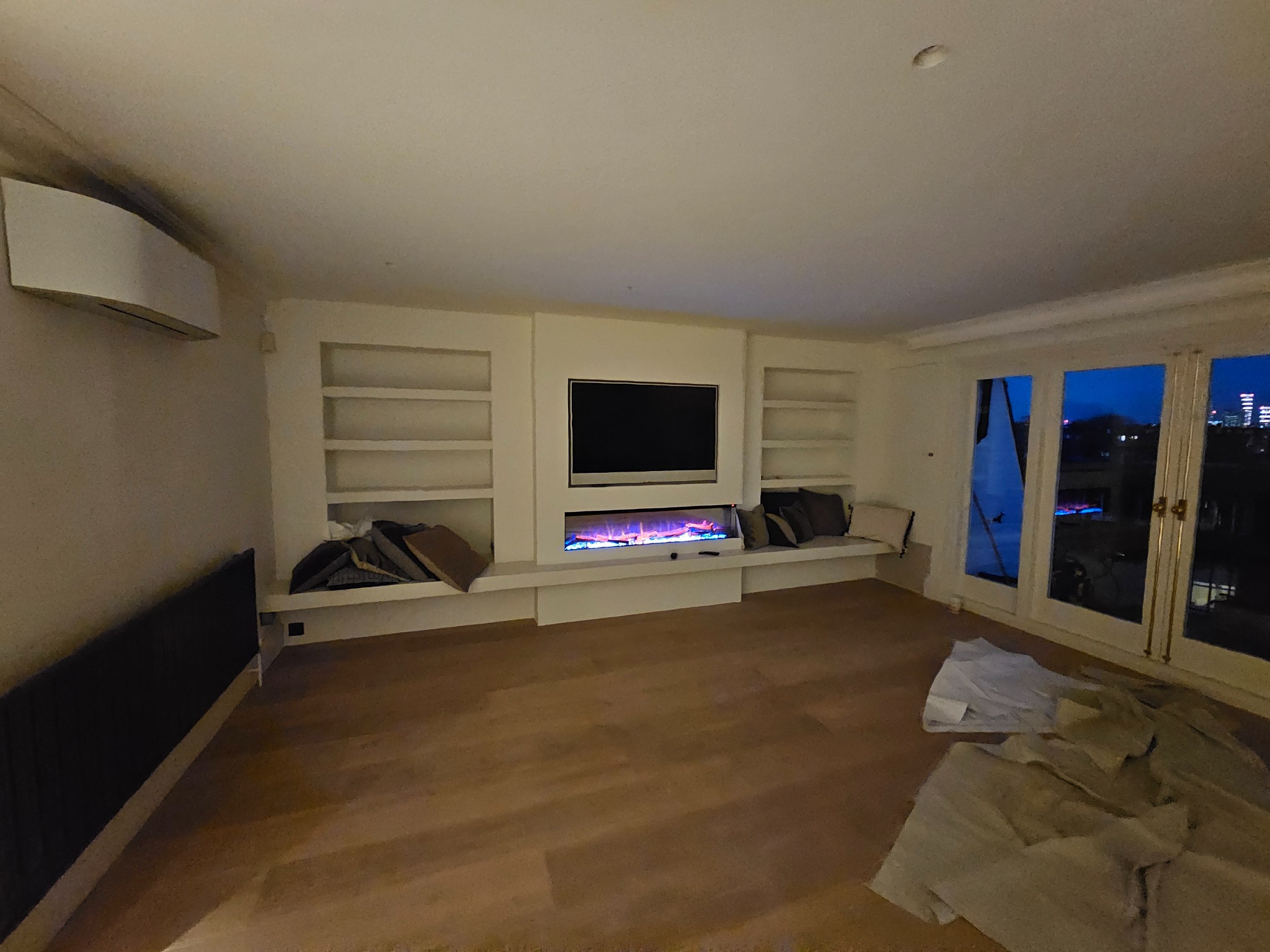 Nowoczesny salon z drewnianą podłogą, zabudową ściany z telewizorem i elektrycznym kominkiem oraz widokiem na nocne miasto przez przeszklone drzwi balkonowe. Część podłogi zabezpieczona folią...