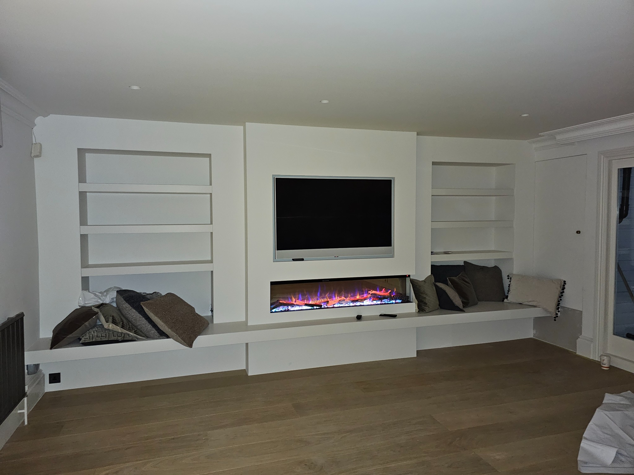 Nowoczesny salon z zabudową meblową na ścianie, wnęki z półkami, telewizor i elektryczny kominek z efektem płomieni, drewniana podłoga