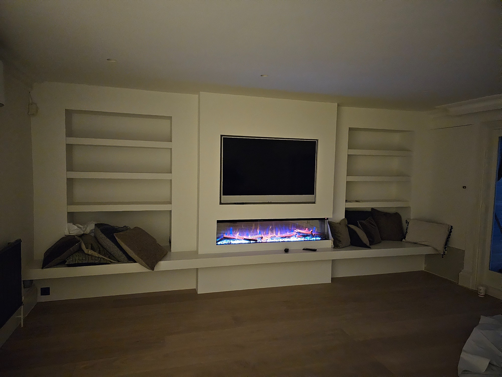 Wnęka w salonie z telewizorem, elektrycznym kominkiem i półkami, oświetlona ciepłym światłem, z drewnianą podłogą.