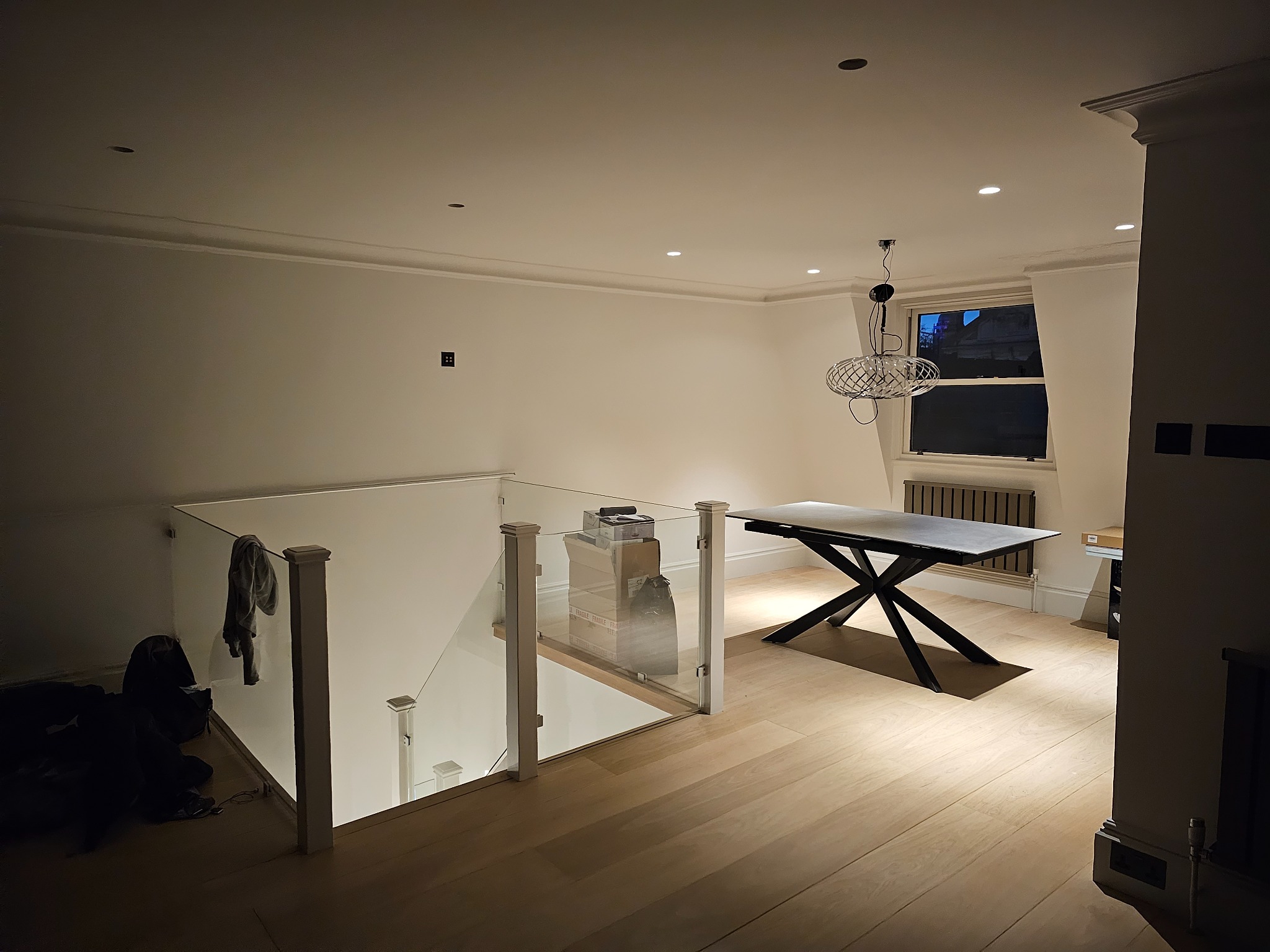 Wnętrze domu po remoncie: jasna podłoga, przeszklona balustrada schodów, nowoczesny stół z czarną podstawą, minimalistyczne oświetlenie sufitowe i dekoracyjna lampa wisząca.