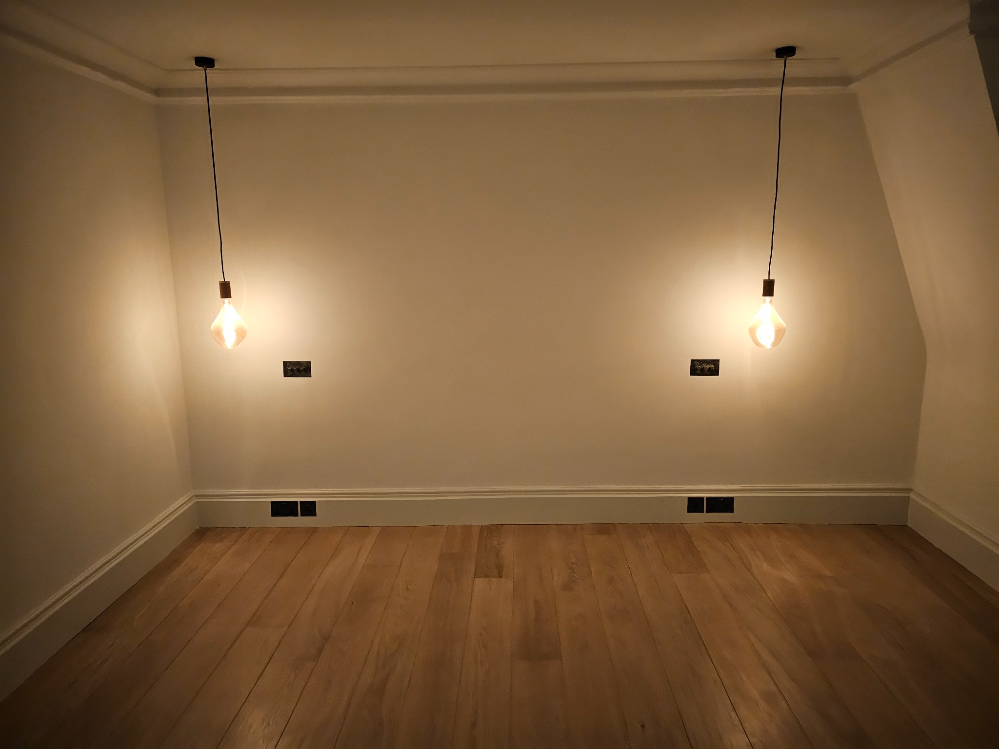 Puste pomieszczenie z jasnymi ścianami, drewnianą podłogą i dwoma wiszącymi żarówkami w stylu retro, oświetlającymi przestrzeń.