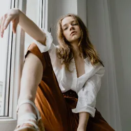 Portret kobiety siedzącej na parapecie w białej koszuli i brązowych spodniach, spoglądającej w dół, z nogą w sandałach na platformie wysuniętą do przodu.