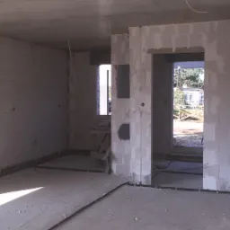 Surowy stan wnętrza domu w trakcie budowy z widocznymi przewodami elektrycznymi ułożonymi wzdłuż ścian i podłogi, oraz puszkami instalacyjnymi w ścianach.