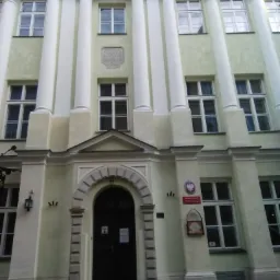 Fasada odnowionego budynku z kolumnami, tablicą pamiątkową i godłem państwowym, widoczne okna z białymi ramami i ciemne drzwi wejściowe.