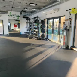 Wnętrze siłowni z profesjonalnym sprzętem do ćwiczeń siłowych, widoczne stojaki na obciążenia, maszyna do ćwiczeń nóg marki KESER, gumowa podłoga i duże okna z widokiem na zewnątrz.