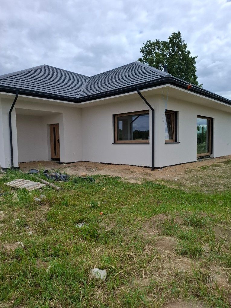 Parterowy dom z szarą dachówką i jasną elewacją, widok z zewnątrz na tle zielonego trawnika i pochmurnego nieba. Widoczne okna z drewnianą ramą i czarne rynny.