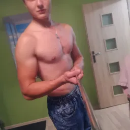 Mężczyzna z odsłoniętym torsem robiący sobie selfie w dżinsach, widoczny tatuaż na ramieniu, w tle zielona ściana i jasne drzwi.