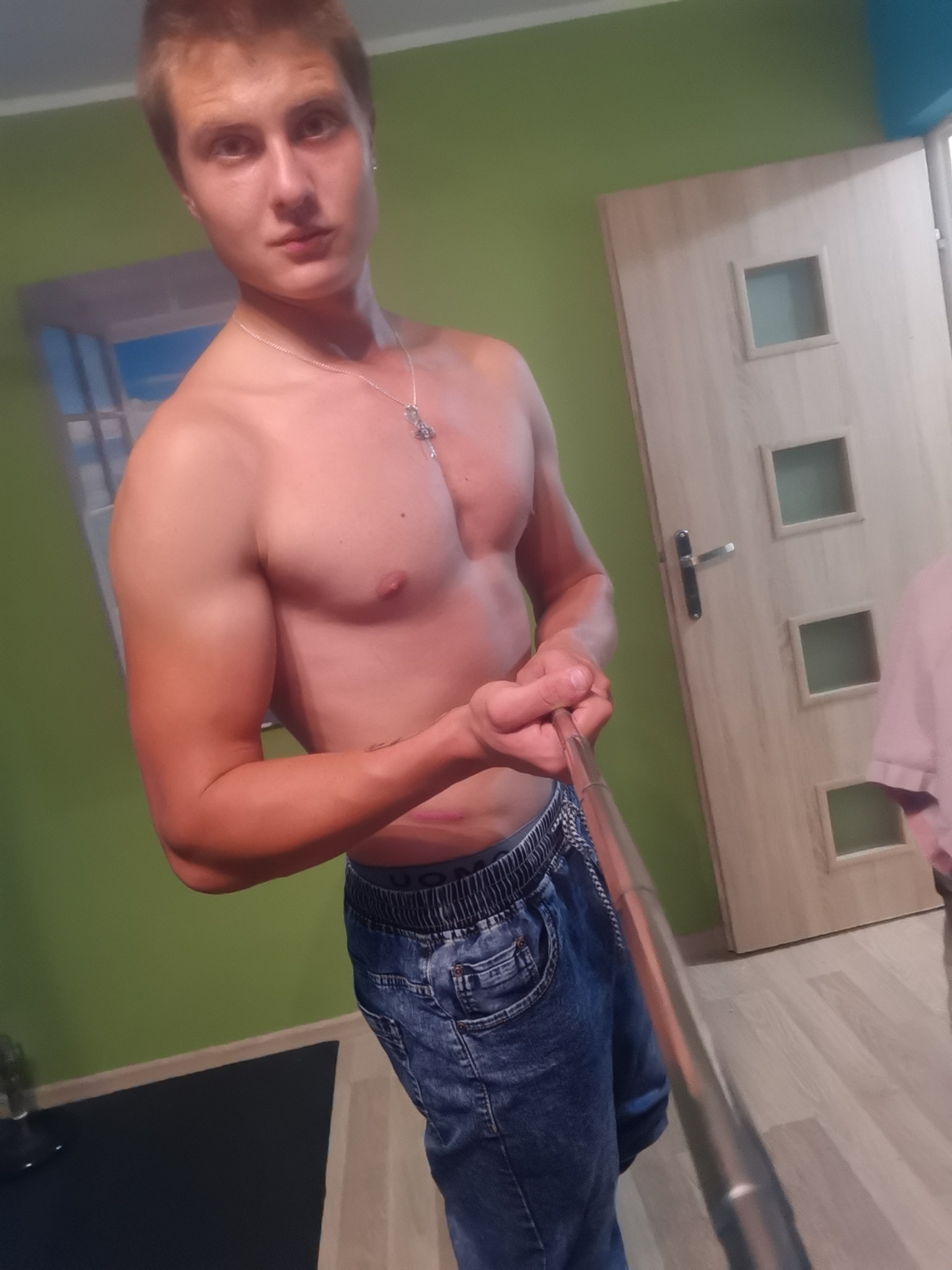 Mężczyzna z odsłoniętym torsem robiący sobie selfie w dżinsach, widoczny tatuaż na ramieniu, w tle zielona ściana i jasne drzwi.