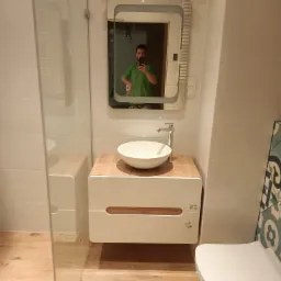 Nowoczesna łazienka z kabiną prysznicową ze szkła, wiszącą szafką z umywalką nablatową i lustrem z podświetleniem LED, odbicie fotografa w lustrze.