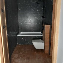 DREWREM USŁUGI REMONTOWO-STOLARSKIE RAFAŁ CHYLA - Nowoczesna łazienka z czarnymi płytkami imitującymi marmur, drewnopodobną podłogą i białym sanitariatem, widziana zza drewnianej ościeżnicy.