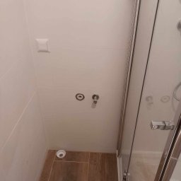 DREWREM USŁUGI REMONTOWO-STOLARSKIE RAFAŁ CHYLA - Wykończona kabina prysznicowa z białymi płytkami ściennymi i drewnopodobną podłogą. Widoczne przyłącza wodne i odpływ. Minimalistyczny styl.