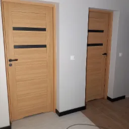 Dwa nowoczesne, drewniane drzwi z czarnymi akcentami w jasnym wnętrzu. Drzwi po lewej stronie z klamką, widoczne listwy przypodłogowe i fragment podłogi.