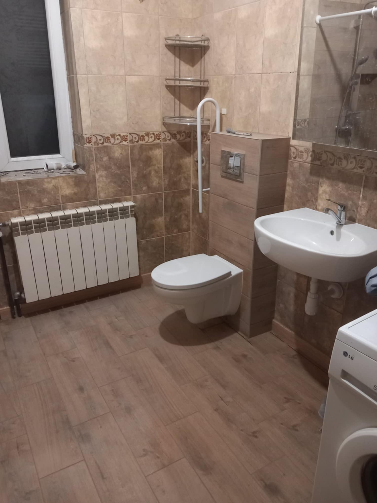 Wykończona łazienka z beżowymi płytkami, białą ceramiką, grzejnikiem i pralką. Widok na umywalkę, toaletę i fragment kabiny prysznicowej.