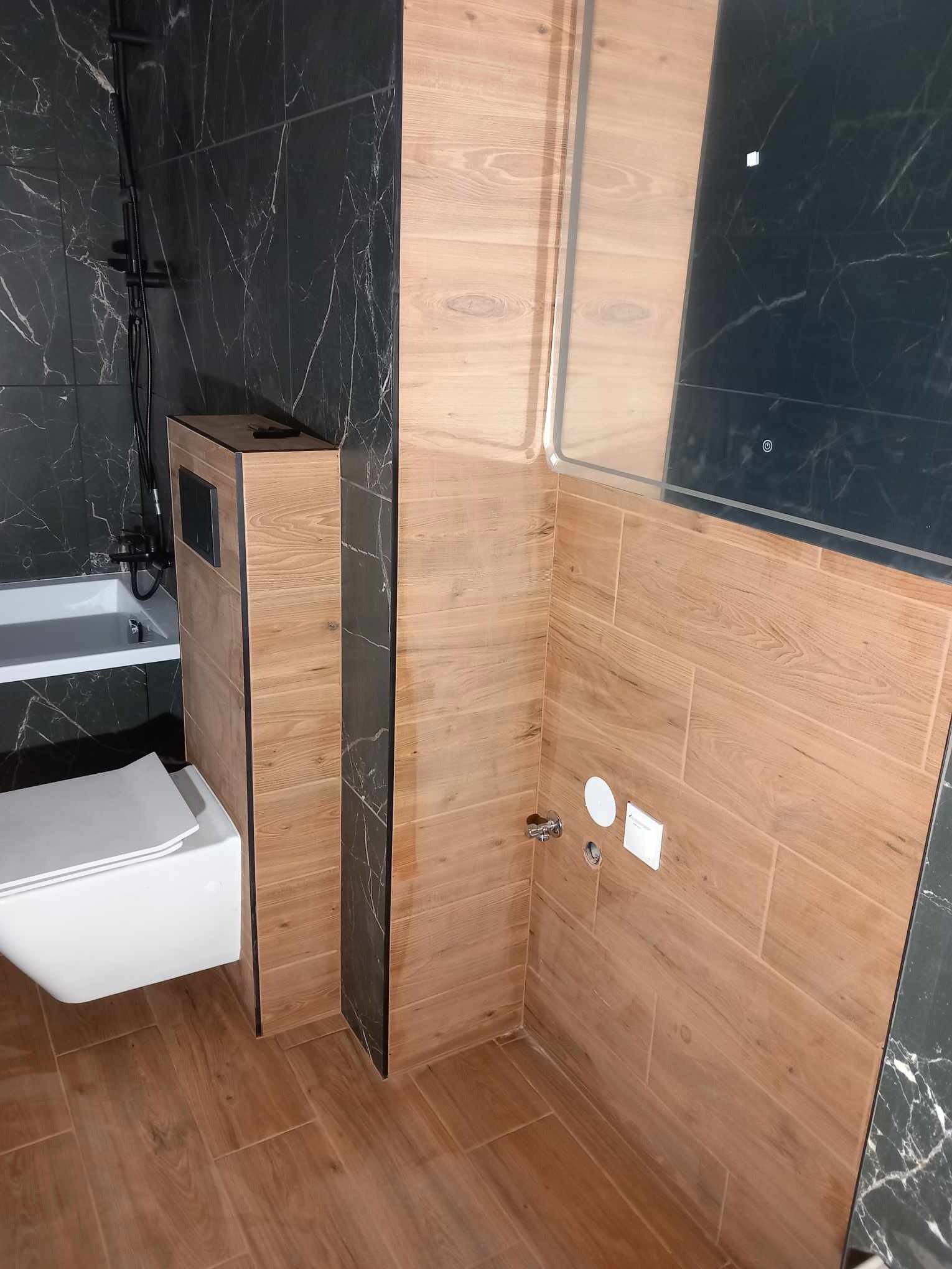 Nowoczesna łazienka z drewnopodobnymi i marmurowymi płytkami, wisząca toaleta i zabudowa geberit z elementami czerni. Minimalistyczny design i funkcjonalność.