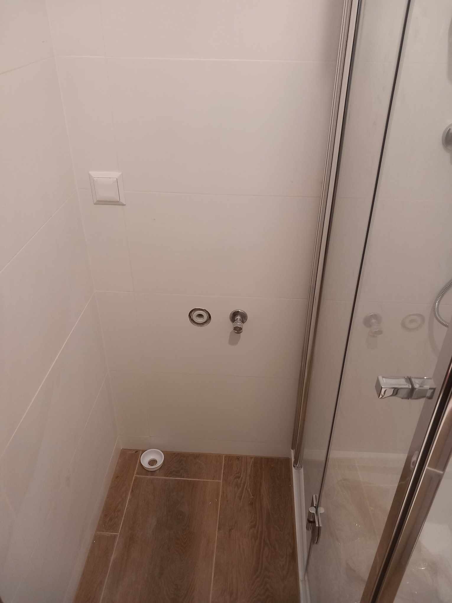 Wykończona kabina prysznicowa z białymi płytkami ściennymi i drewnopodobną podłogą. Widoczne przyłącza wodne i odpływ. Minimalistyczny styl.