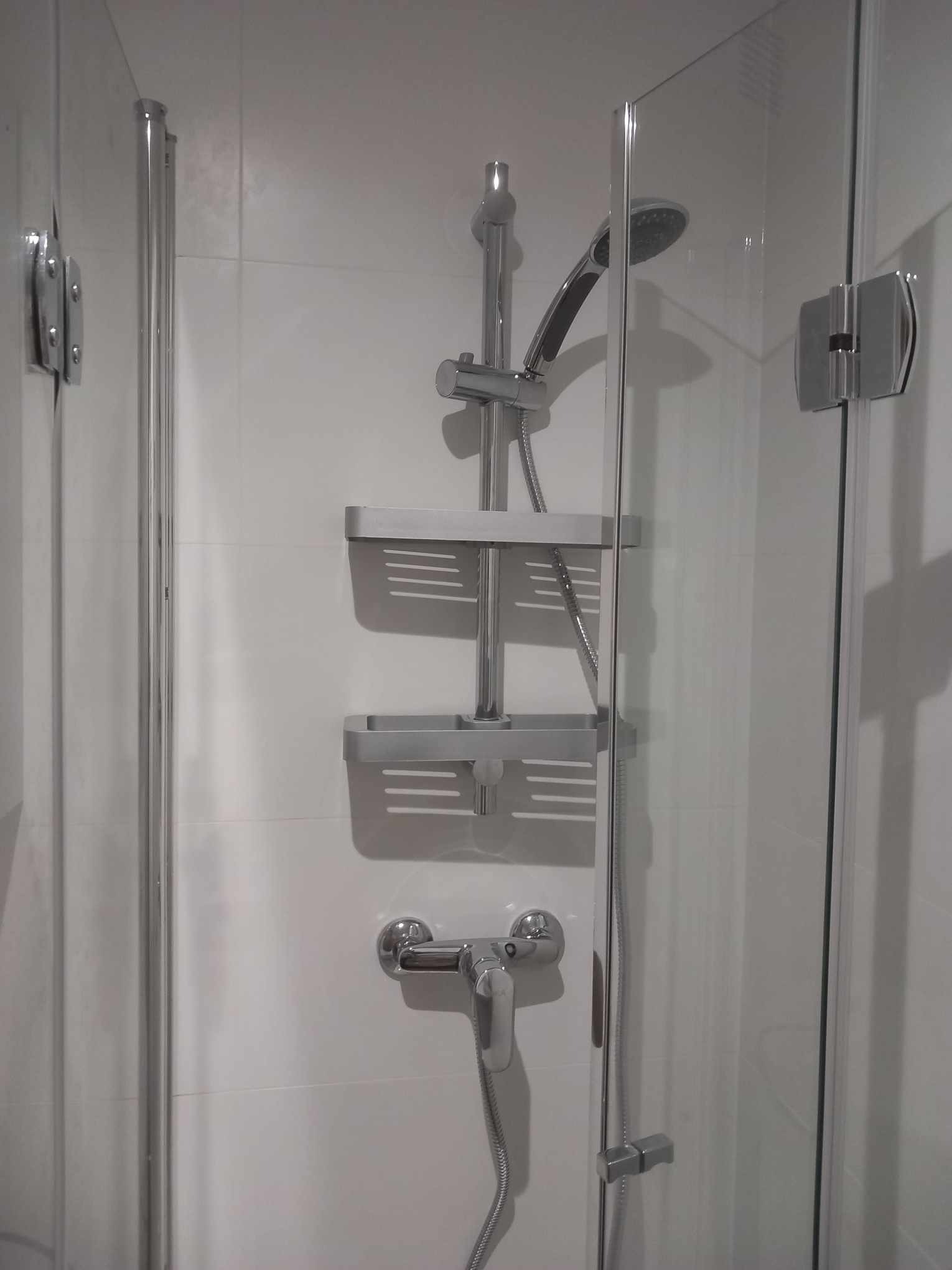 Nowoczesna kabina prysznicowa z chromowaną armaturą i półkami na kosmetyki, białe płytki na ścianach, minimalistyczny design.