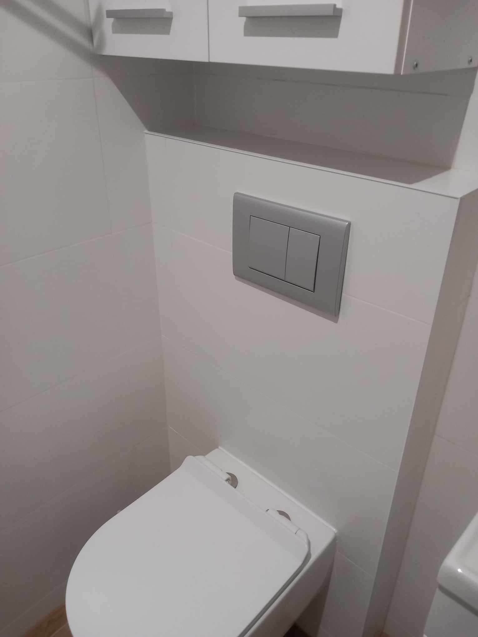 Nowoczesna łazienka: biała toaleta podwieszana z szarym przyciskiem spłukiwania, białe płytki i szafka wisząca. Minimalistyczny design.