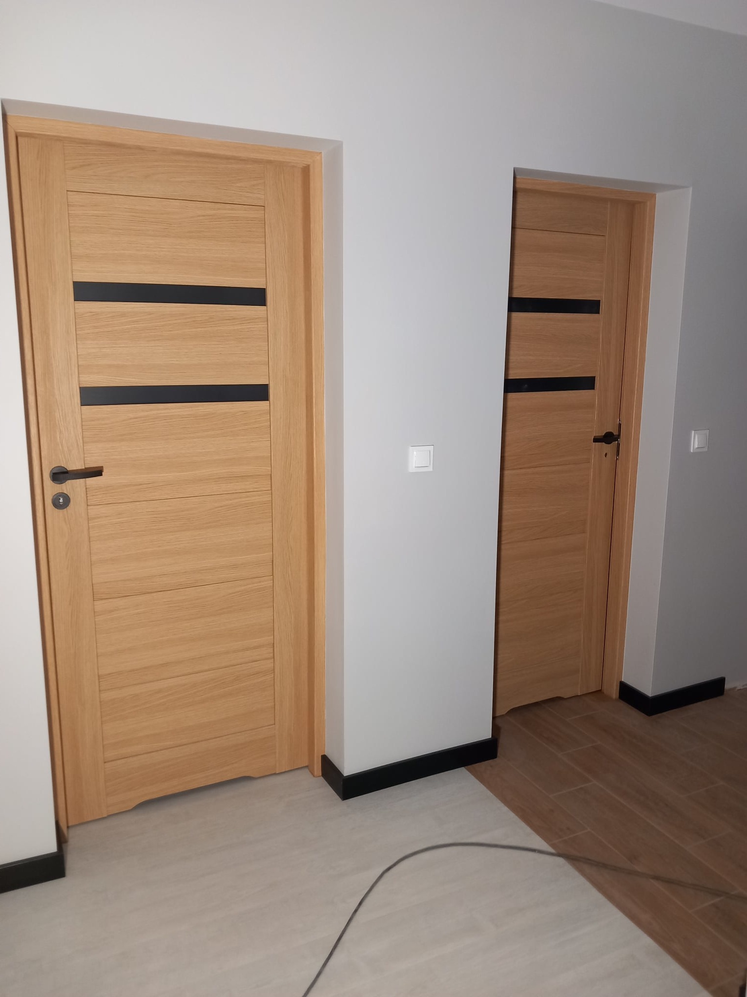 Dwa nowoczesne, drewniane drzwi z czarnymi akcentami w jasnym wnętrzu. Drzwi po lewej stronie z klamką, widoczne listwy przypodłogowe i fragment podłogi.