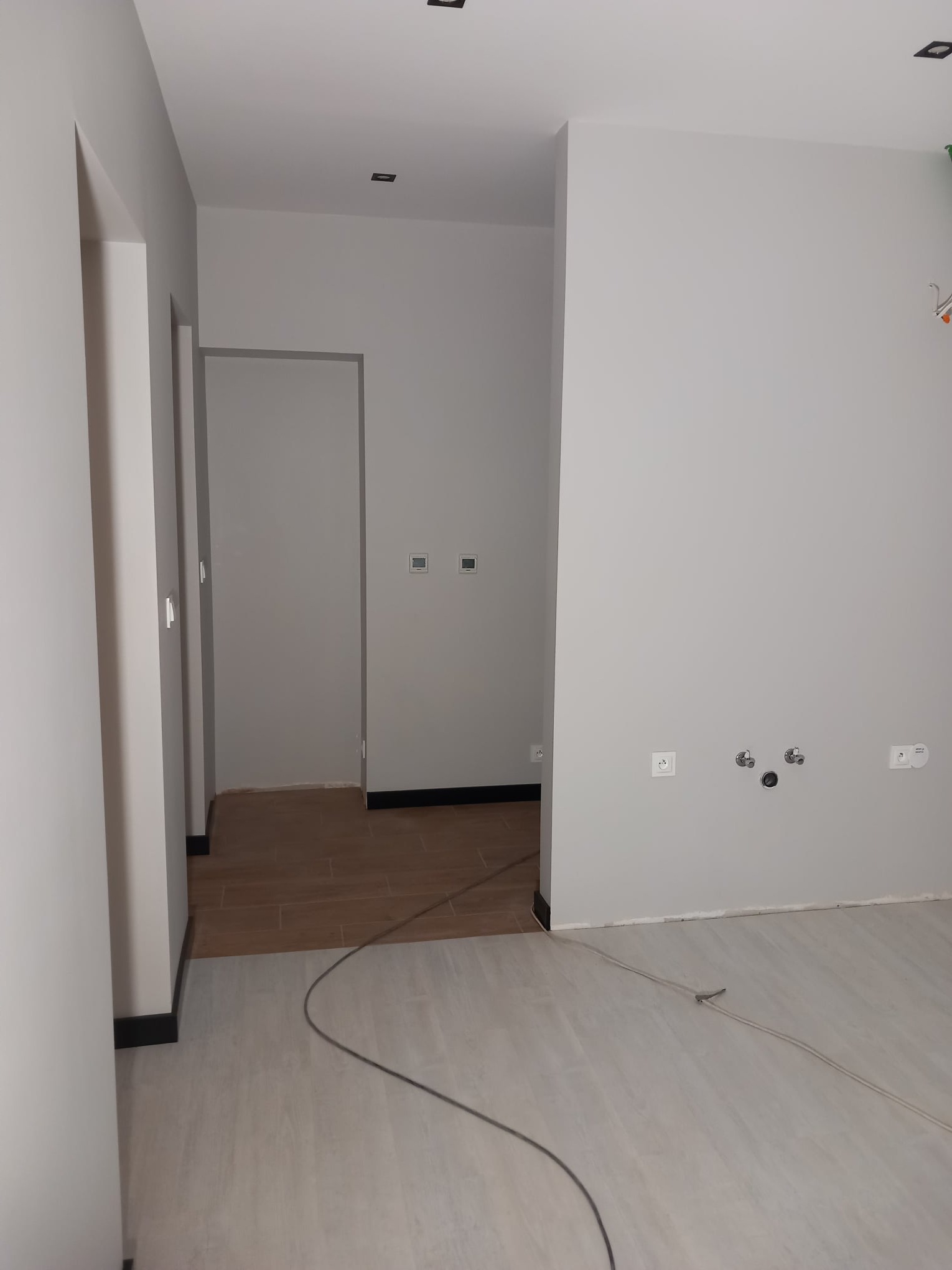 Jasne, świeżo pomalowane wnętrze w odcieniach szarości. Widoczne gniazdka elektryczne, rury i fragmenty podłogi z paneli oraz płytek. Minimalistyczny design.