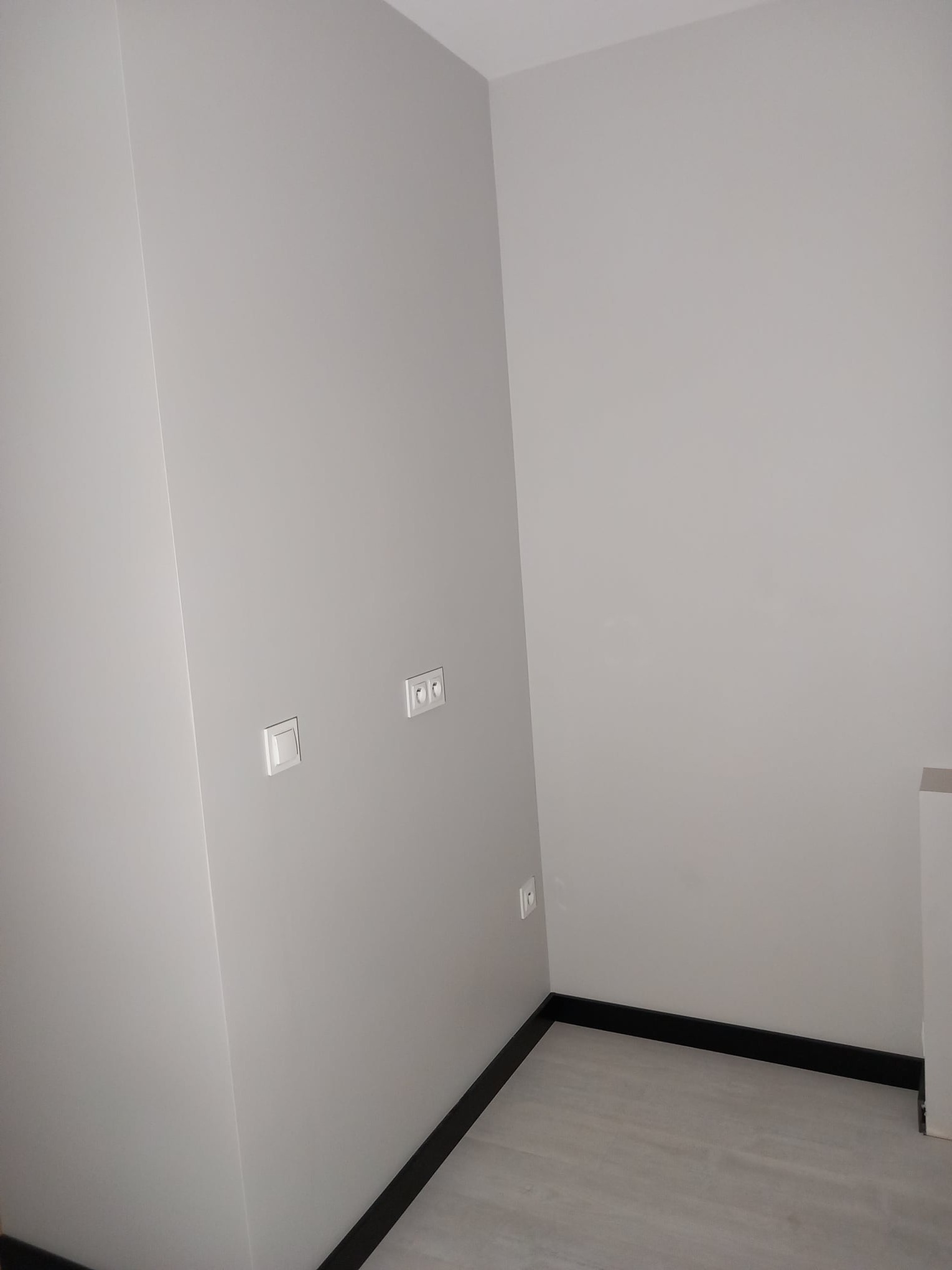 Jasne wnętrze po malowaniu ścian na szaro, widoczne gniazdka elektryczne i czarne listwy przypodłogowe, podłoga wykończona panelami.