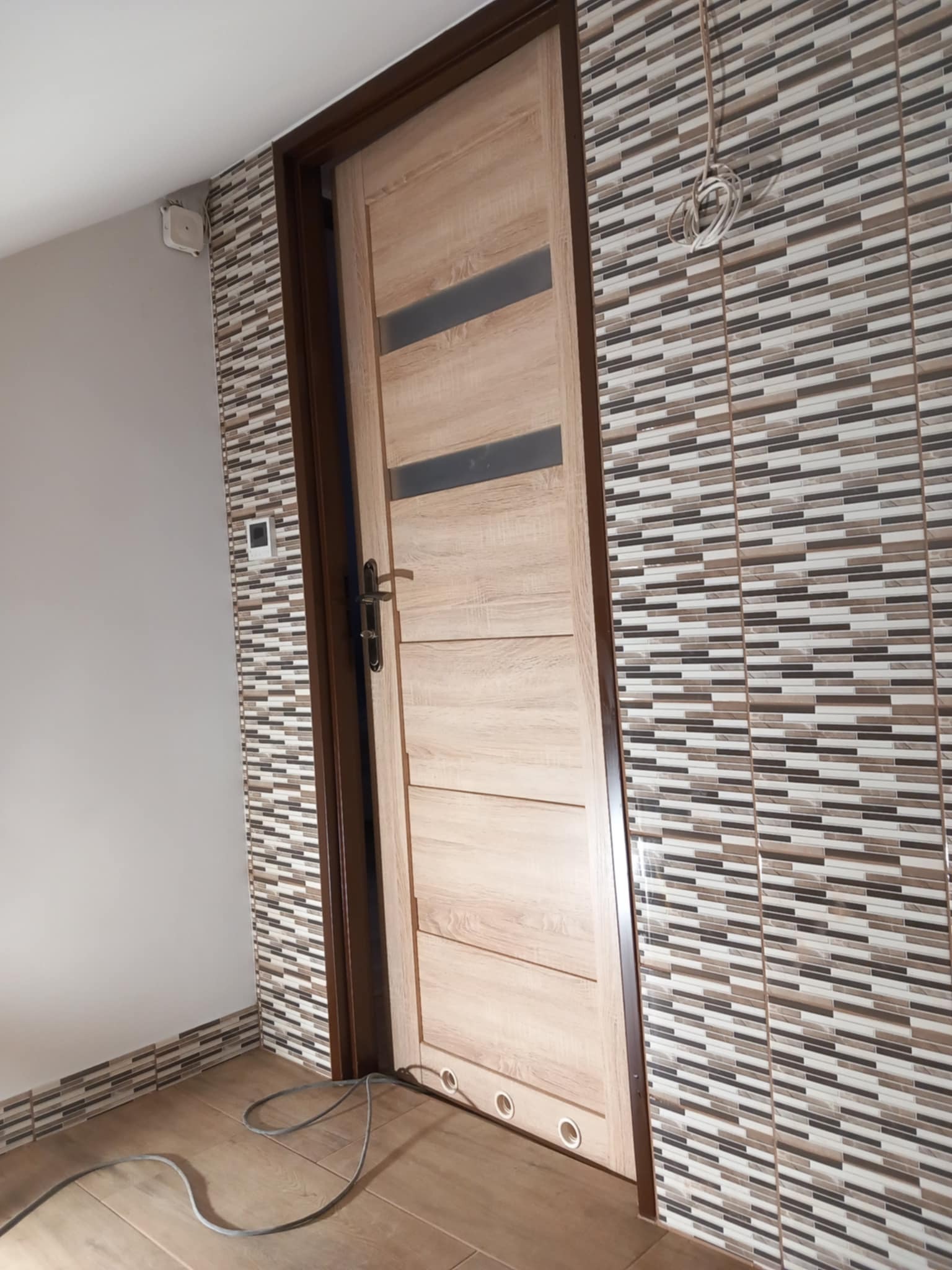 Zamontowane drzwi wewnętrzne z szybami w nowo wykończonym wnętrzu. Na ścianie mozaika z płytek w odcieniach brązu i beżu. Na podłodze panele w ciepłym odcieniu drewna.