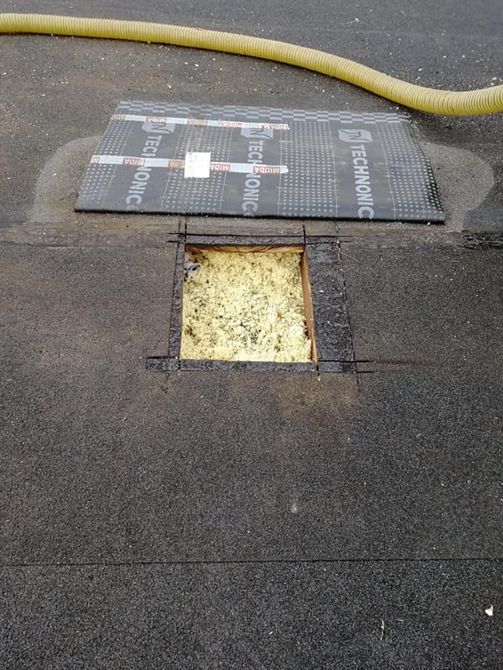 Stropodach z wyciętym otworem ukazującym żółtą pianę izolacyjną, obok leży arkusz papy z nadrukiem Technonicol i żółty wąż do wdmuchiwania izolacji.