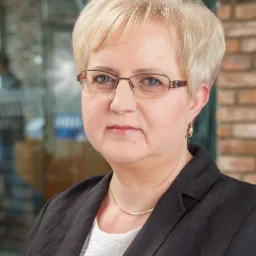 Portret kobiety w okularach i czarnej marynarce, z krótkimi blond włosami, na tle biurowym z ceglaną ścianą i szklaną ścianką działową.