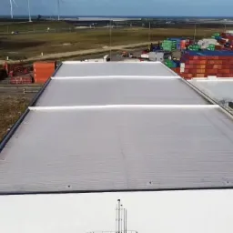 Rozległy, płaski dach przemysłowy z szarej membrany, widoczny z góry, z elementami infrastruktury portowej i turbinami wiatrowymi w tle.