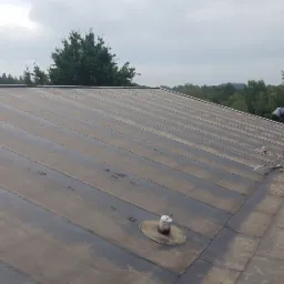 Widok na płaski dach pokryty papą termozgrzewalną podczas prac dekarskich, widoczny dekarz, rolki papy, narzędzia i kask ochronny.