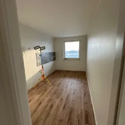 Wnętrze pokoju z drewnopodobną podłogą, świeżo pomalowanymi ścianami i fragmentem dekoracyjnej mozaiki, oświetlone lampą budowlaną na trójnogu.