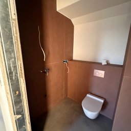 Tomasz Olender usługi remontowo budowlane wykończenia wnętrz - Łazienka w trakcie wykańczania: brązowe płytki ścienne, szara podłoga, wisząca toaleta, instalacja elektryczna i wodna w trakcie montażu, widoczna ościeżnica drzwi.