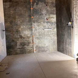 Tomasz Olender usługi remontowo budowlane wykończenia wnętrz - Surowa łazienka w trakcie wykańczania: ciemne, imitujące rdzę płytki na ścianach z pomarańczowymi krzyżykami dystansowymi, jasne płytki podłogowe, widoczne instalacje wodne i elektryczne, surowy...