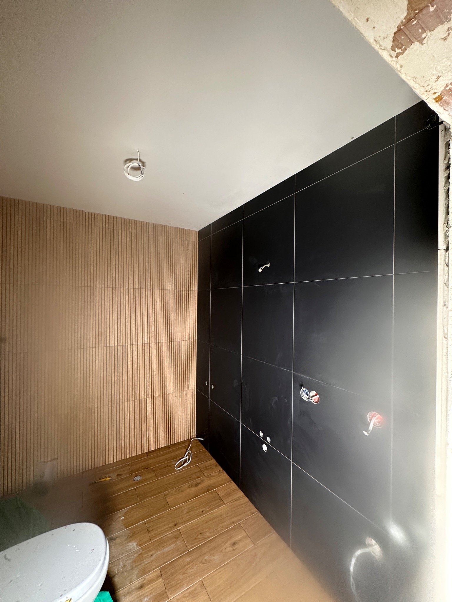 Łazienka w trakcie prac wykończeniowych: czarne, błyszczące płytki ścienne z widocznymi instalacjami, drewnopodobna okładzina ścienna z pionowych paneli, oraz podłoga z paneli imitujących drewno...