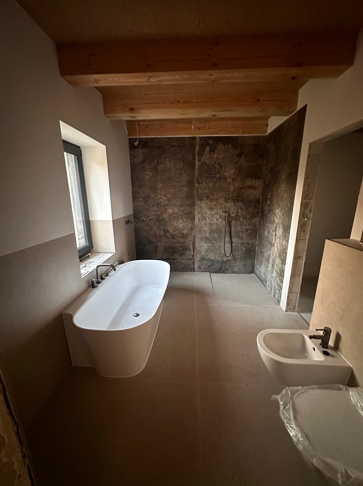 Wnętrze łazienki w trakcie wykańczania: biała wanna wolnostojąca pod oknem, kabina prysznicowa typu walk-in z szarymi płytkami imitującymi beton, bidet, drewniane belki stropowe, częściowo zakryta...