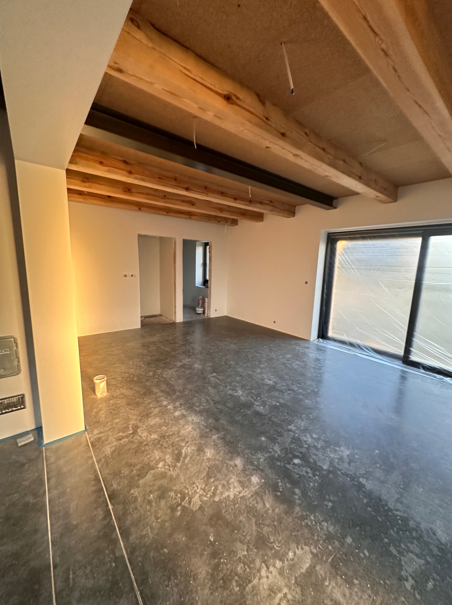 Surowe drewniane belki stropowe w nowo wykończonym wnętrzu, z szarą, błyszczącą posadzką i przygotowanymi do malowania ścianami, okno zabezpieczone folią.