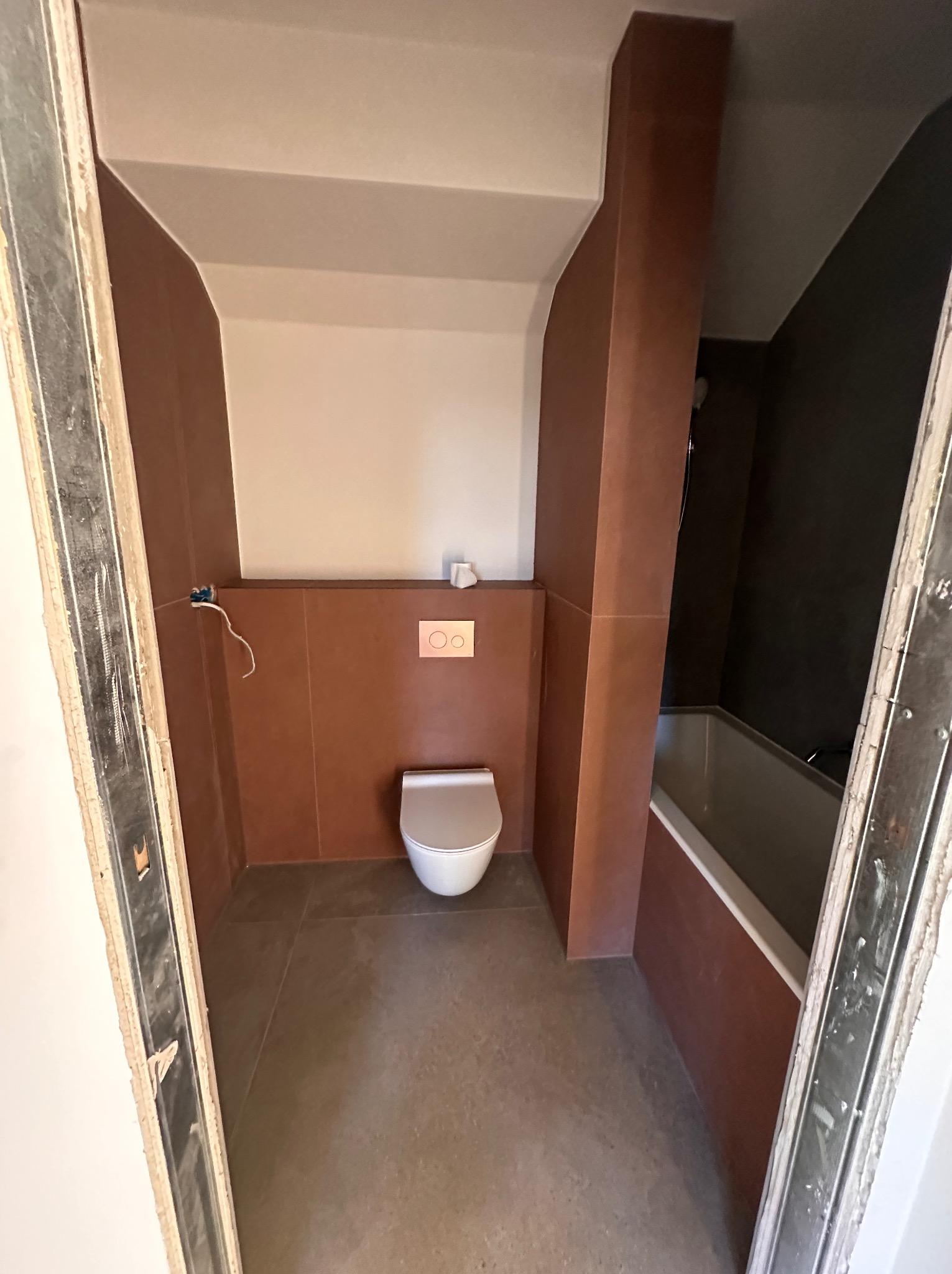 Łazienka w trakcie wykańczania w Mrągowie: widok na częściowo wykończoną toaletę podwieszaną z brązowymi płytkami, widoczne niedokończone obramowanie drzwi i instalacja elektryczna.