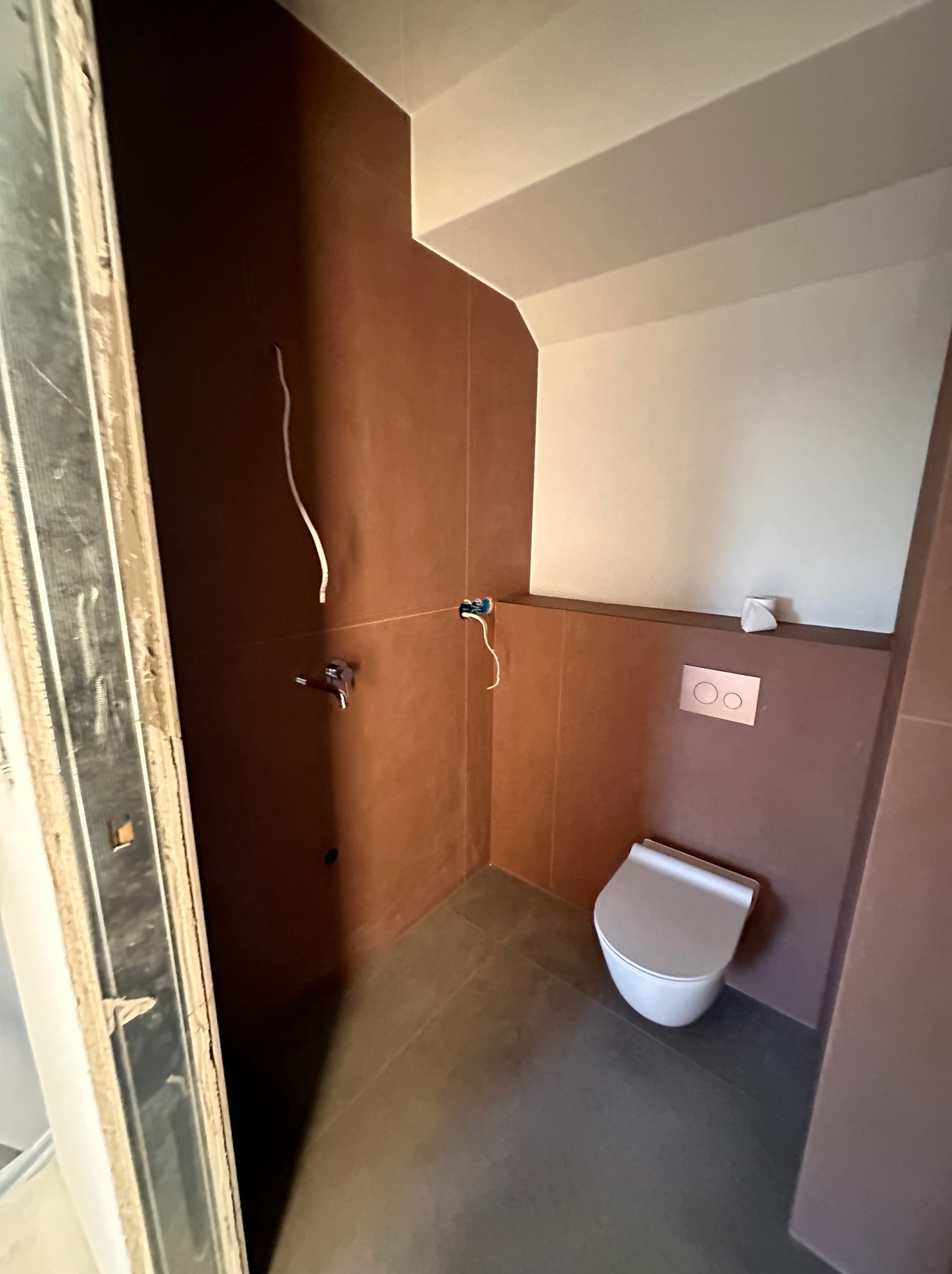 Łazienka w trakcie wykańczania: brązowe płytki ścienne, szara podłoga, wisząca toaleta, instalacja elektryczna i wodna w trakcie montażu, widoczna ościeżnica drzwi.