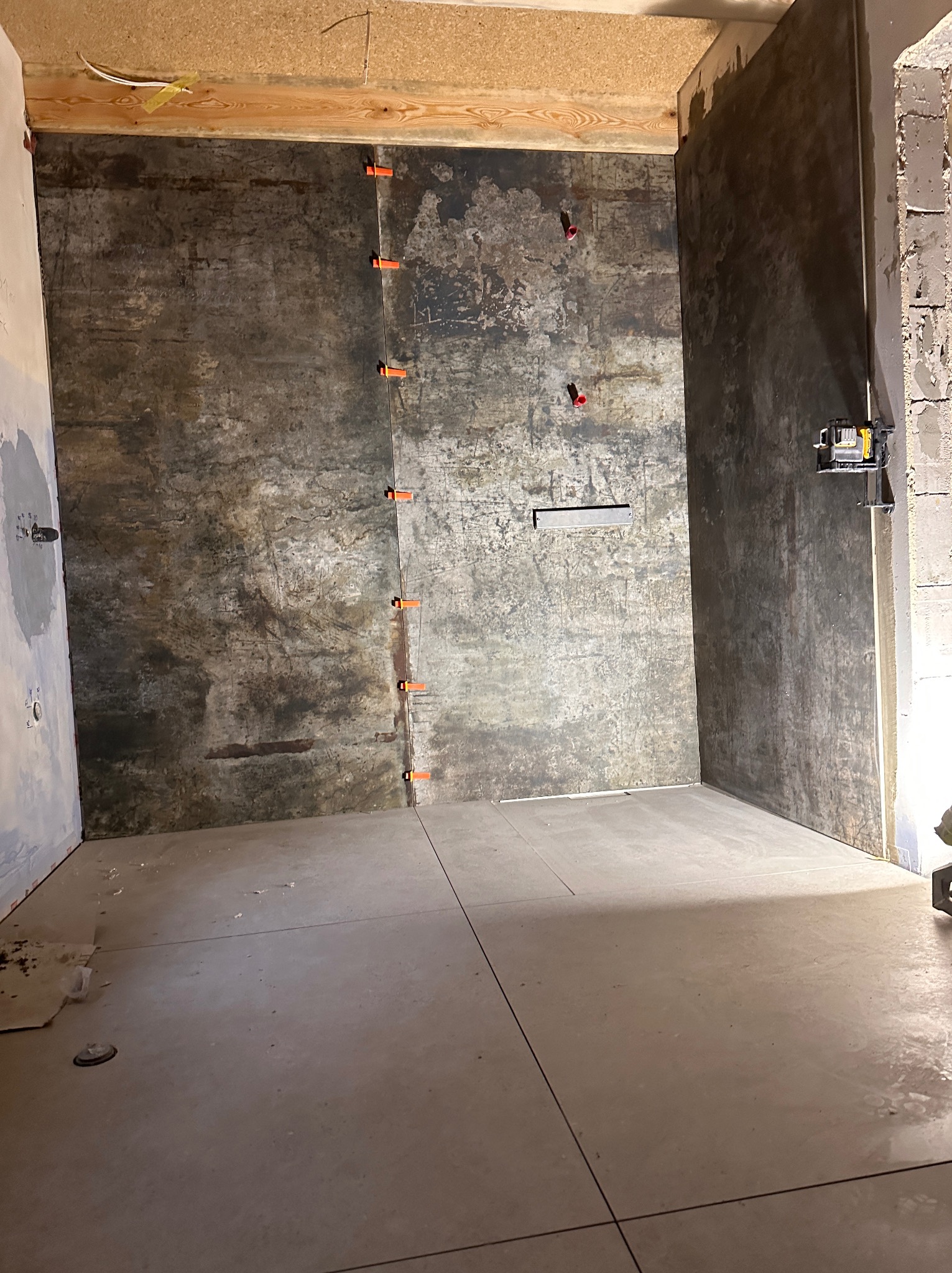 Surowa łazienka w trakcie wykańczania: ciemne, imitujące rdzę płytki na ścianach z pomarańczowymi krzyżykami dystansowymi, jasne płytki podłogowe, widoczne instalacje wodne i elektryczne, surowy...