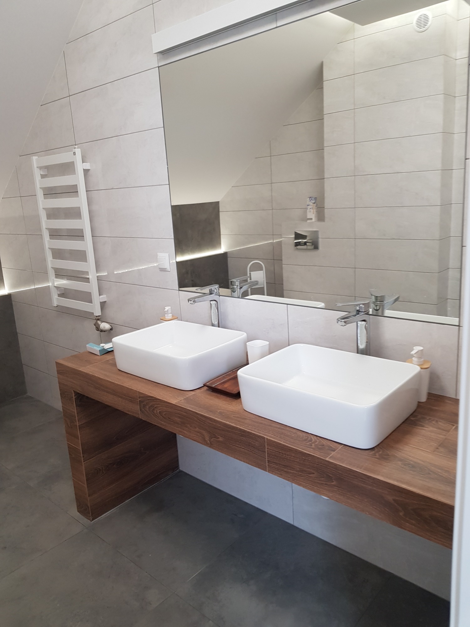 Nowoczesna łazienka w Przemyślu z dwiema kwadratowymi umywalkami nablatowymi na drewnianym blacie, duże lustro, grzejnik drabinkowy i szare płytki ścienne.