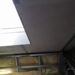 Niedokończona konstrukcja sufitu z metalowych profili i fragmentów płyt, widoczna izolacja z wełny mineralnej w ścianie, ujęcie z dołu pod kątem, oświetlenie dzienne.