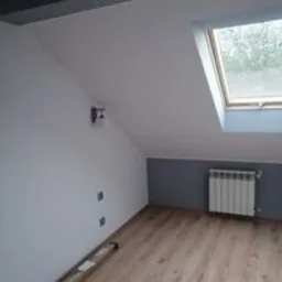 Poddasze po remoncie: białe ściany, drewniana podłoga, grzejnik pod skosem, okno dachowe z widokiem na zewnątrz, szary pas pomalowany na dolnej części ściany.