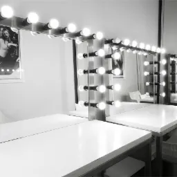 Czarno-białe zdjęcie pustej garderoby z lustrami otoczonymi żarówkami, białe blaty i designerskie krzesła, na ścianie plakat z modelką.