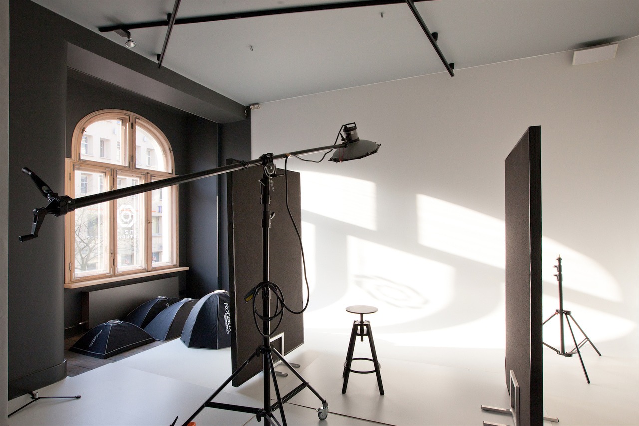 Studio fotograficzne z oknem łukowym, lampą na wysięgniku, blendami i stołkiem w minimalistycznym wnętrzu z białym tłem.