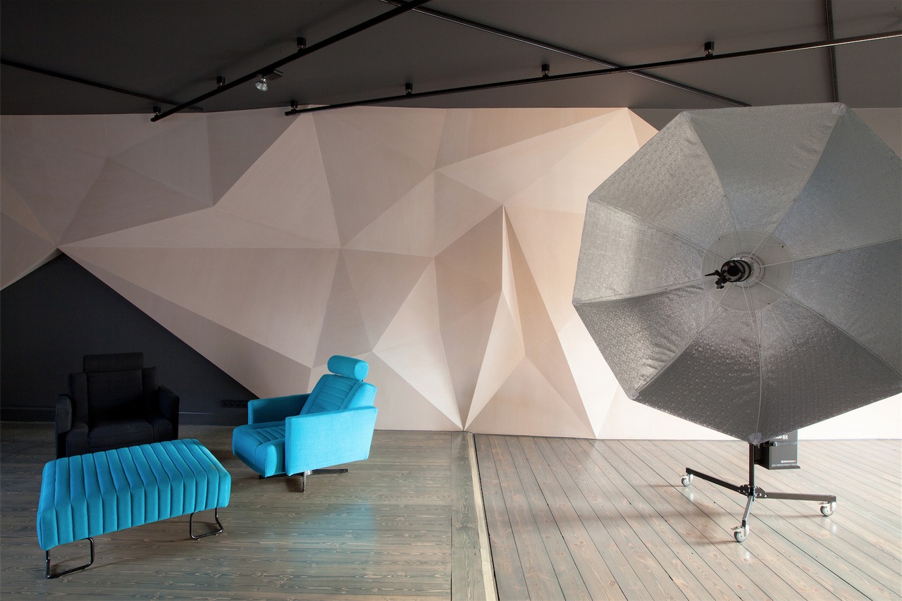 Studio fotograficzne z geometryczną ścianą, turkusowym fotelem i pufą oraz czarnym fotelem, oświetlone lampą studyjną z softboxem.