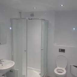 Nowoczesna łazienka z kabiną prysznicową narożną, białą umywalką z szafką i toaletą podwieszaną, oświetlona punktowymi lampami sufitowymi.