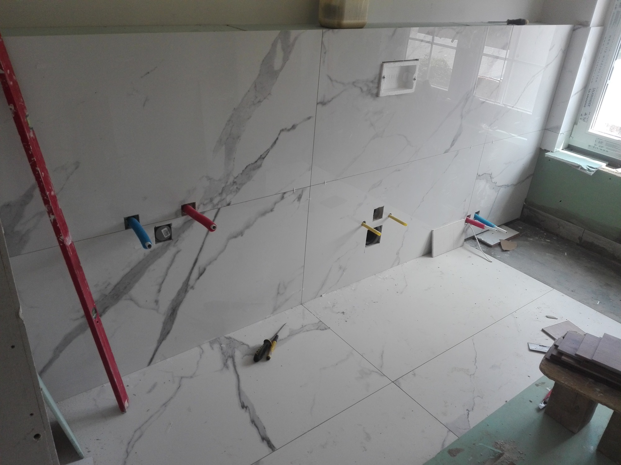 Ściana i podłoga w trakcie układania płytek imitujących marmur, widoczne wystające rury instalacji wodnej i poziomica oparta o ścianę.